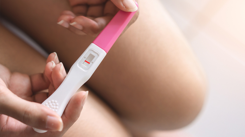 A person holds a white pregnancy test with a pink cap, showing a single red line in the result window, positioned against their lap in soft lighting.